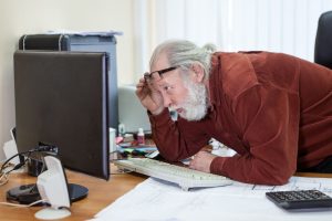 Retirement office employee looks at the monitor screen, lifting his glasses on his forehead and moving closer